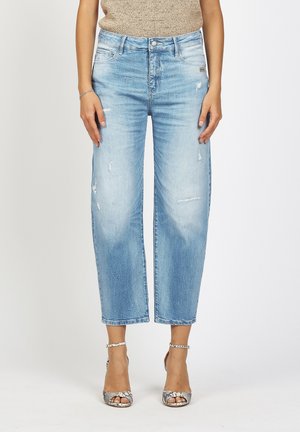 Frau trägt hellblaue, leicht ausgefranste Cropped-Jeans, ein beige ärmelloses Oberteil und Schlangenhaut-Ankle-Strap-Absätze, steht vor weißem Hintergrund.