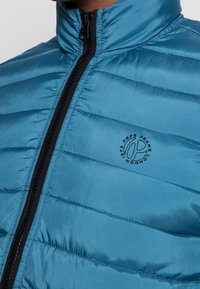 Detailní záběr na osobu, která má na sobě modrou prošívanou bundu s černým zipem a logem Pepe Jeans London na hrudi.
