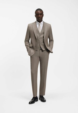 Man draagt een beige driedelig pak met wit overhemd, bijpassende stropdas, zwarte nette schoenen, staand met handen in de zakken op een witte achtergrond.
