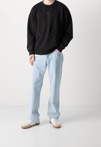 Persona che indossa una felpa nera a maniche lunghe, jeans azzurri dal taglio comodo e sneakers bianche, in piedi davanti a uno sfondo semplice.