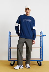 Navy blue sweatshirt with white accents and "Puma" logo, olive green pants, and white athletic shoes with purple detailing. Blue cart in the background.