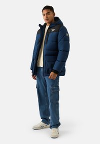 Giacca imbottita blu navy con pannello nero sulla spalla e logo Reebok. Abbinata a jeans cargo azzurri e sneakers bianche. Tessuto strutturato.
