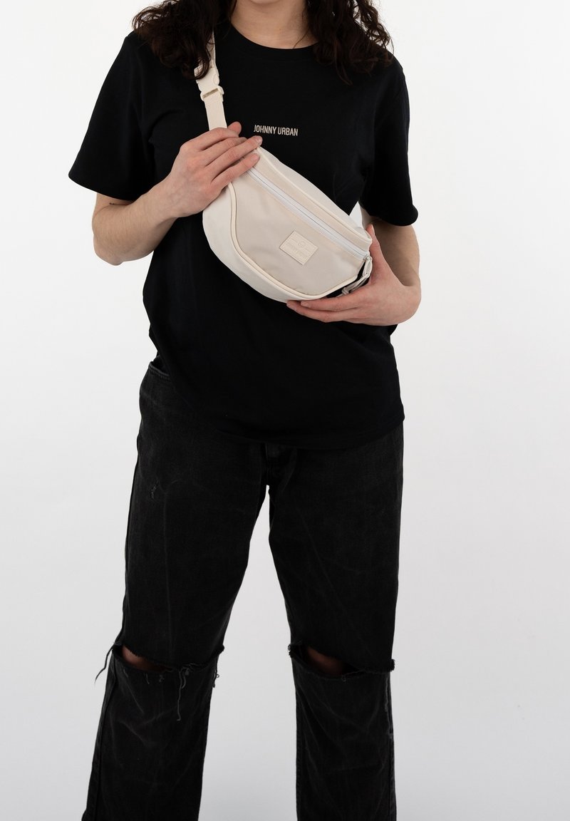 Personne portant un jean noir déchiré et un t-shirt noir tenant un sac bandoulière beige sur un fond blanc.