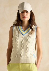 Kreminė kabelio pynimo liemenė su mėlynu ir geltonu juostuotu V formos apykakle, turinti priglundantį briaunotą hemą ir mažą mėlyną logotipo detalę.