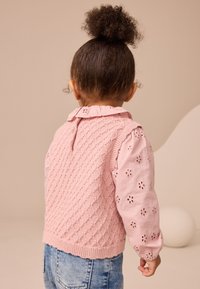 Lyserød strikket vest med et tekstureret diamantmønster, kombineret med en langærmet lyserød bluse med blomsterdetaljer i hulmønster.