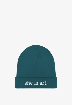 Bonnet tricoté bleu sarcelle avec un revers sur lequel est brodée en blanc la phrase « she is art. ». Texture lisse, épaisseur moyenne.