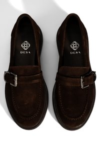 Braune Wildlederslipper mit einer Schnallendetail, runder Spitze und einer strukturierten schwarzen Sohle. Logo auf der Innensohle sichtbar. Schlichtes und elegantes Design.