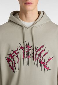 Beige hoodie met een vooraanstaande grafiek met stekelige zwarte en roze letters. Materiaal lijkt zacht met een gezellige textuur; inclusief trekkoorden.