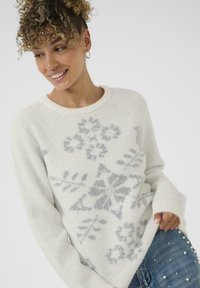 Pull en tricot blanc avec un motif de flocon de neige argenté, col et poignets côtelés, et texture douce. Associé à un jean bleu avec des accents de perles.