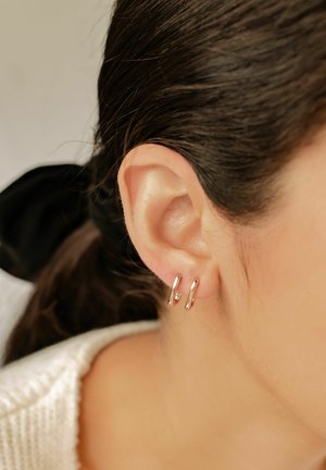 Gros plan sur l'oreille d'une femme avec deux petites boucles d'oreilles en or en forme d'anneau, des cheveux bruns attachés en arrière avec un chouchou noir, et un vêtement en tricot blanc.