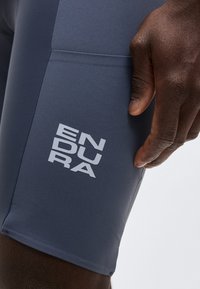 Χέρι με σκουρόχρωμο δέρμα ακουμπισμένο δίπλα σε σκούρο μπλε σορτς με λευκό λογότυπο "ENDURA" στον μηρό, πάνω σε απλό ανοιχτό φόντο.