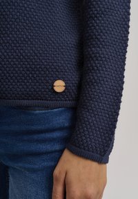 Navyblauer Strickpullover mit strukturiertem Muster, runder Holzlogo-Akzent nahe dem Saum und gerippten Bündchen. Kombiniert mit Denim-Stoff.