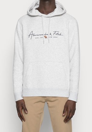 Mørkhudet person iført lysegrå Abercrombie & Fitch hættetrøje med logo og beige bukser, stående mod ensfarvet baggrund.
