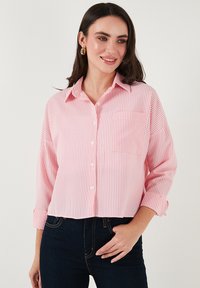 Pink and white striped button-up shirt with a front pocket, cropped cut, and rolled sleeves, paired with dark jeans.