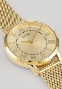 Reloj de pulsera dorado con correa de malla metálica, caja redonda, marcadores de horas en números romanos y una esfera texturizada dorada. Nombre de la marca "GUESS" mostrado.