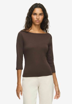 Femme aux cheveux bouclés et foncés, portant un haut marron ajusté à manches trois-quarts et un pantalon clair, debout devant un fond uni.