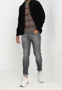 Schwarze Cordhemd, gemusterter grau-roter Pullover, graue Jeans und weiße Sneaker mit farbigen Akzenten. Lässiges Outfit mit unterschiedlichen Texturen.