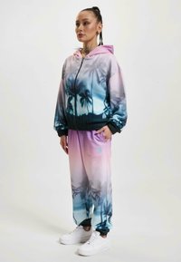Completo sportivo con una gradazione di colori rosa e blu e stampa di palme; giacca con zip e cappuccio, polsini elasticizzati e pantaloni abbinati.