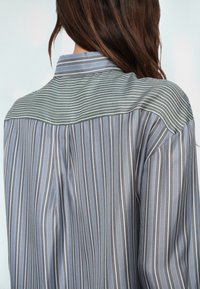 Femme aux longs cheveux bruns portant une chemise rayée grise et bleue avec un empiècement et un col rayés verts et blancs contrastants, vue de dos.