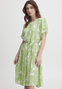 Rochie verde cu mâneci scurte, cu un model abstract alb, talie strânsă și fustă vaporoasă. Fabricată dintr-un material ușor, cu o textură moale.