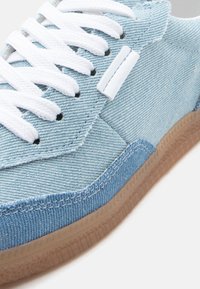 Ανοιχτό μπλε τζιν αθλητικό παπούτσι με λευκά κορδόνια και σόλα από καουτσούκ gum, εμφανιζόμενο σε κοντινή λήψη υπό γωνία.