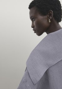 Profil latéral d'une personne aux cheveux courts et bouclés portant des boucles d'oreilles créoles dorées et un vêtement rayé bleu et blanc sur un fond uni.