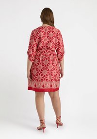 Red patterned dress with a tie belt, three-quarter sleeves, and a scalloped hem. Features intricate floral designs in gold. Paired with red heels.