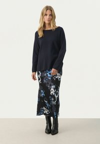 Pull marin avec un décolleté large, associé à une jupe à motif floral aux tons bleu et noir, complétée par des bottes à talons noirs.