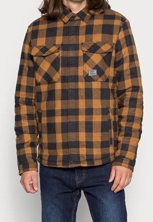 Mann trägt ein braun-schwarz kariertes Hemd mit Knöpfen und zwei Brusttaschen sowie dunkelblaue Jeans, steht vor einem einfarbigen Hintergrund.