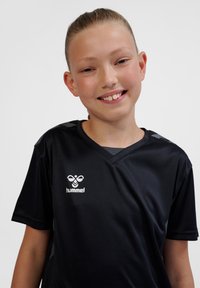 Lachende jonge persoon met naar achteren getrokken haar, gekleed in een zwart hummel sportshirt tegen een effen witte achtergrond.