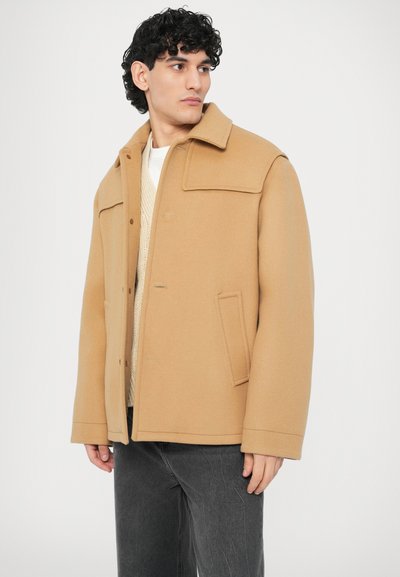 Καμηλό παλτό από μείγμα μαλλιού με δομημένο γιακά, κλείσιμο με κουμπιά μπροστά, δύο πλαϊνές τσέπες και άνετη γραμμή, φορεμένο πάνω από μπεζ πλεκτό πουλόβερ.