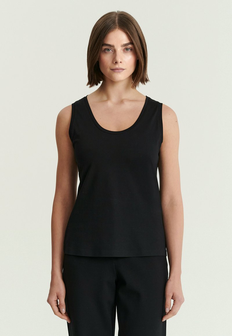 Femme aux cheveux courts et bruns, vêtue d'un haut noir sans manches et d’un pantalon noir, debout devant un fond clair uni.