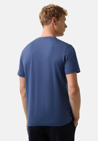 Mand med kort, krøllet blond hår iført en ensfarvet blå t-shirt med korte ærmer, stående med ryggen til og hænderne i lommerne.
