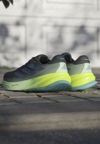 Zwart en grijs sportieve schoenen met drie blauwe strepen, mesh bovenkant en felgroene tussenzool. Beschikt over een gestructureerd ontwerp en een contrasterende zool.
