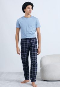 Tricou din bumbac albastru deschis, cu mâneci scurte, asortat cu pantaloni de pijama în carouri bleumarin. Modelul stă desculț pe o suprafață moale, gri.