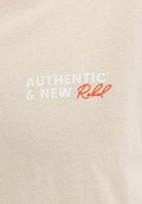 Tissu beige avec le texte "AUTHENTIC & NEW" en lettres majuscules blanches et "Rebel" en écriture cursive rouge.