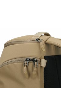 Esquina de una mochila de tela beige que muestra dos compartimentos con cremallera y tiradores color canela, además de un panel trasero acolchado negro.