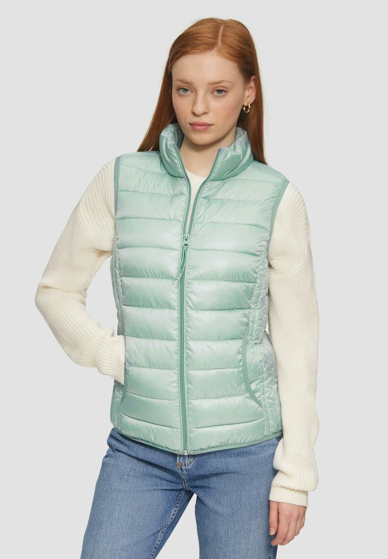Hellgrüne gepolsterte Weste mit hohem Kragen, Reißverschluss vorne und zwei Seitentaschen. Hat eine glatte Textur und ein gestepptes Design. Kombiniert mit einem cremefarbenen Sweater.