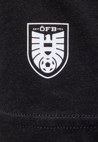 ÖFB-logo i hvidt på sort stof, med en stiliseret ørn over en fodbold inde i et skjold med teksten "SEIT 1904".