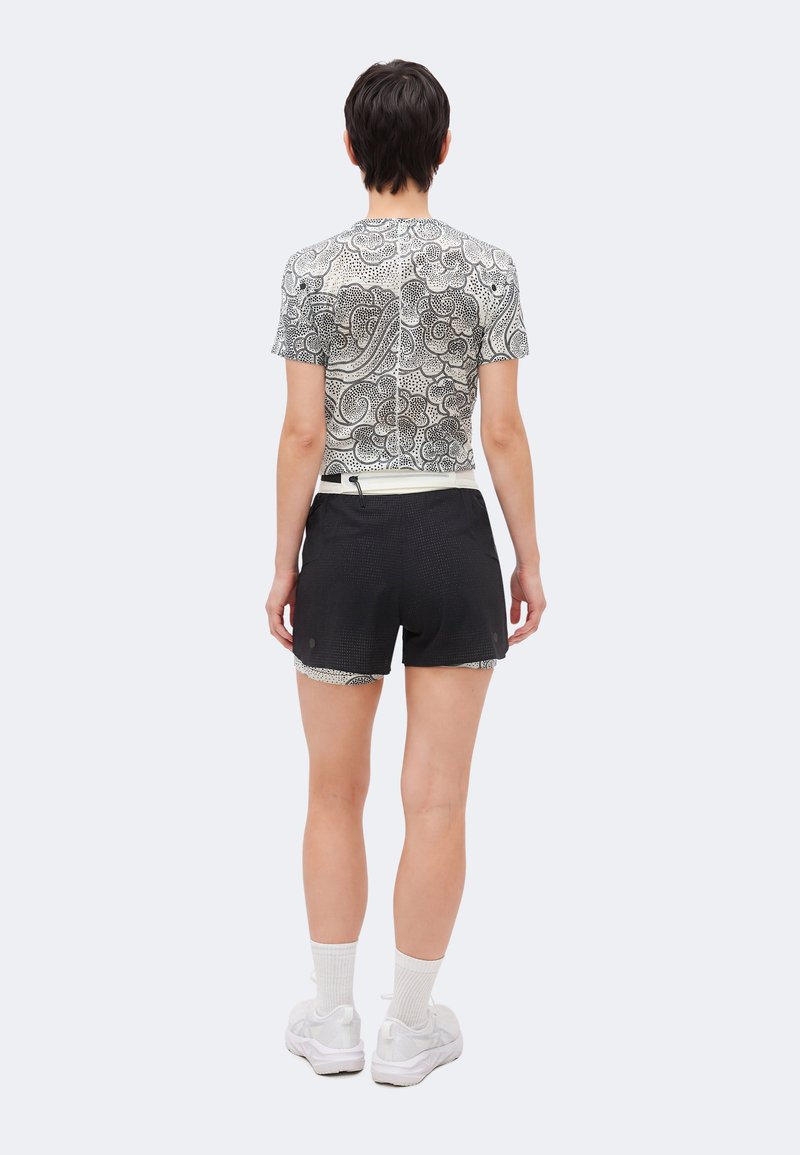 Person, der står med ryggen til iført en mønstret hvid og sort kortærmet skjorte, sorte shorts med hvid kant, hvide sokker og hvide sneakers.