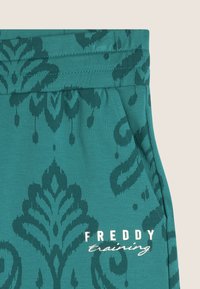 Pantalones de entrenamiento en color verde azulado, hechos de tejido suave, con un estampado floral oscuro, cinturilla elástica y un bolsillo frontal. Logo impreso en el costado.