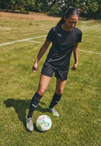 Black soccer jersey and shorts, paired with black and green socks and gray cleats. Features a white logo and vibrant multi-colored soccer ball.