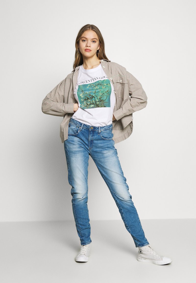 Giovane donna in posa con le mani sui fianchi, che indossa una maglietta bianca con grafica, una camicia overshirt a quadri beige, jeans blu e sneaker alte bianche.