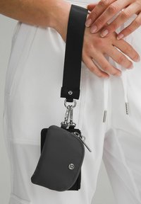 Hands holding a black strap attached to small black zippered pouches hanging from white pants with drawstrings.