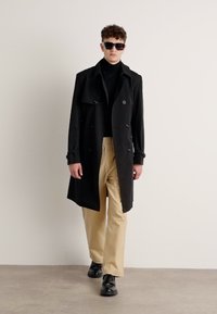 Manteau trench noir à double boutonnage, en tissu léger, superposé sur un pull col roulé noir, associé à un pantalon beige et des chaussures noires.