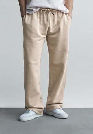 Personne portant un pantalon beige à cordon, un T-shirt blanc et des baskets blanches, debout les mains dans les poches devant un fond uni.