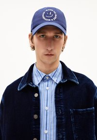 Marineblaue Cordjacke mit silbernen Knöpfen, getragen über einem hellblau gestreiften Hemd, gekrönt von einer blauen Mütze mit einem runden Logo.