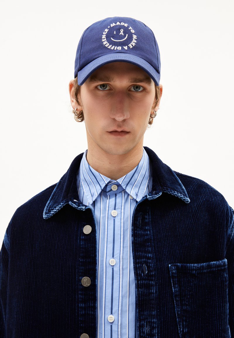 Marineblaue Cordjacke mit silbernen Knöpfen, getragen über einem hellblau gestreiften Hemd, gekrönt von einer blauen Mütze mit einem runden Logo.