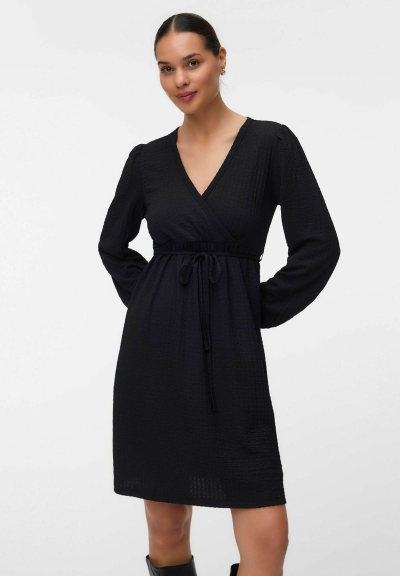 Femme aux cheveux foncés attachés porte une robe portefeuille noire à manches longues avec une ceinture à la taille et des bottes noires, debout sur un fond blanc uni.