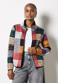 Kobieta w złotych kolczykach w kształcie kół, wielokolorowej kurtce z zamkiem błyskawicznym w stylu patchwork, białej koszuli i szarych dżinsach z wysokim stanem, stoi na jednolitym tle.